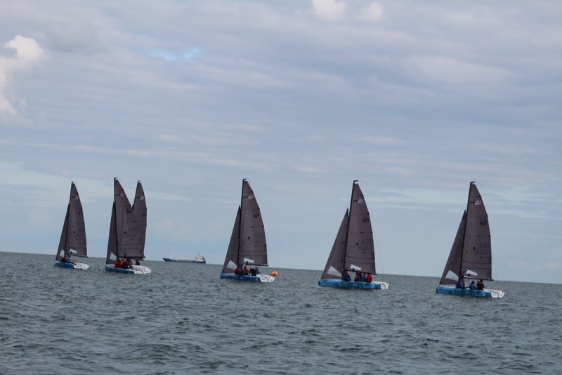 Six boats from our fleet spread along a flat horizon under high cloud, a coaster passing behind them and an orange racing mark to one side.