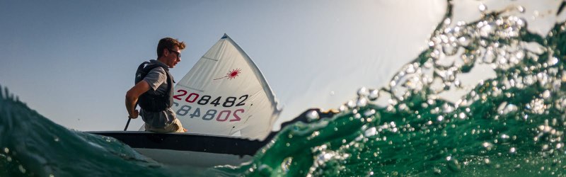 Me sailing 208482, seen from the water through the crest of a breaking wave, sitting up on the side deck with the tiller extension in hand against a clear sky.