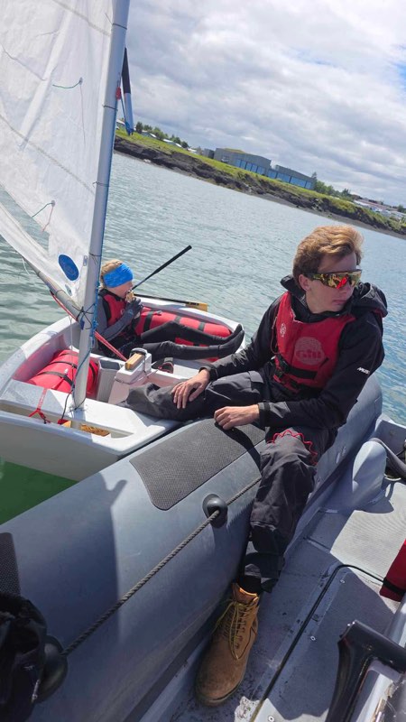 Sitting on the tube of the RIB holding an Optimist alongside between races, the young sailor aboard in a blue headband eating a snack, low Icelandic shoreline and a long grey building behind.