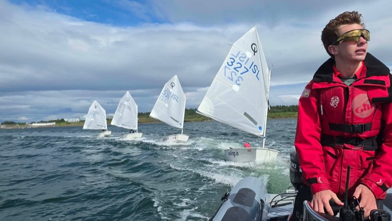 At the console of the RIB in a red drysuit, hand on the throttle, with the Optimists strung out astern under tow, their sails up and ISL numbers showing, low green Icelandic coast behind.