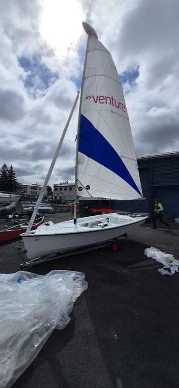 A brand new RS Venture standing on its trolley in the boat park with main and jib hoisted for the first time, the sun burning through broken cloud behind the rig and the packaging still spread on the tarmac.