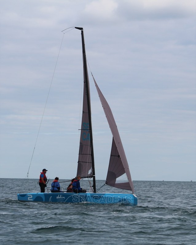 Us running downwind on hull 2 in light air, the mainsail up and the jib hanging soft, all of us gathered in the cockpit under a pale sky.