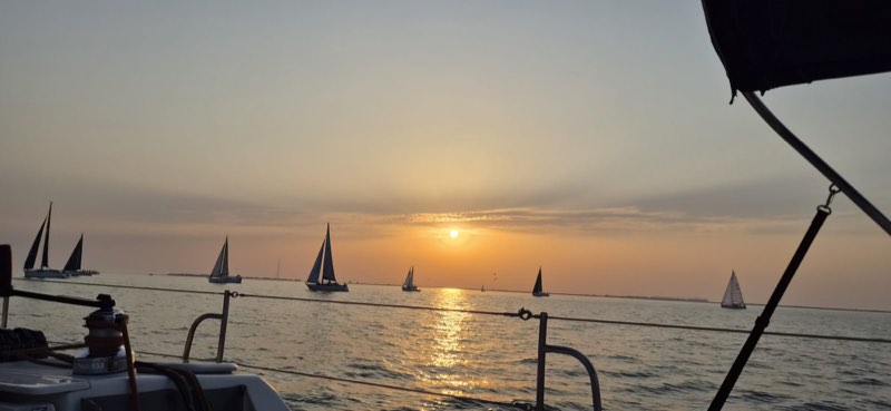 The view out over a yacht guardrail at sunset, eight keelboats spread along the horizon with the sun low and its light laid across the water.
