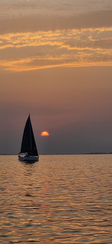 A yacht under main and headsail silhouetted against a low red sun, banded orange cloud above and the last of the light running across flat water toward the camera.